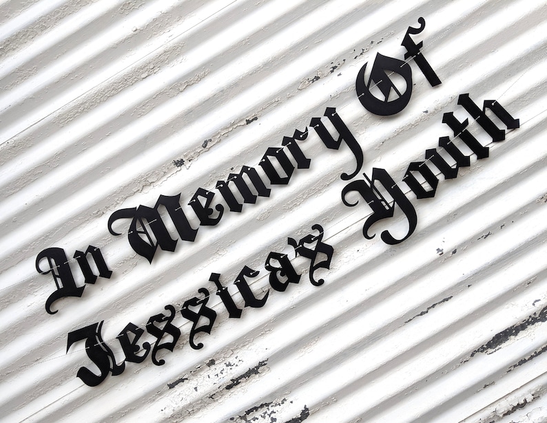 May include: Black metal sign with the words "In Memory Of Jessicaz Youth" in a gothic font. The sign is mounted on a white, weathered, slatted wooden background.