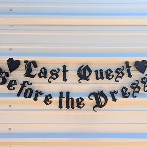 May include: Black metal banner with the text "Last Quest Before the Dress" in a gothic font, strung between two black heart shapes. The banner is displayed against a white and blue striped background.
