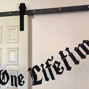 May include: A black paper banner featuring the text "One Love One Lifetime" in a gothic-style font, strung together on a thin white cord. The banner includes a black heart-shaped cutout at the end and is displayed against a white wall.