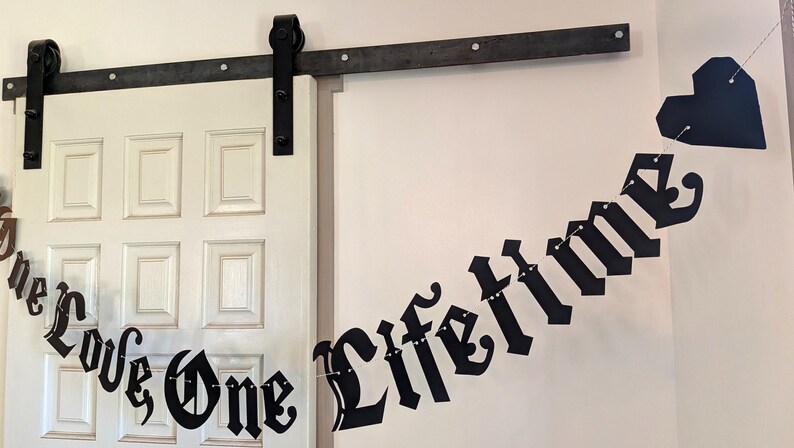 May include: A black paper banner featuring the text "One Love One Lifetime" in a gothic-style font, strung together on a thin white cord. The banner includes a black heart-shaped cutout at the end and is displayed against a white wall.