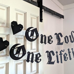 May include: A black paper banner featuring the text One Love One Lifetime in a gothic-style font, interspersed with black heart-shaped cutouts. The individual letters and heart shapes are connected by a thin white string, creating a hanging display.
