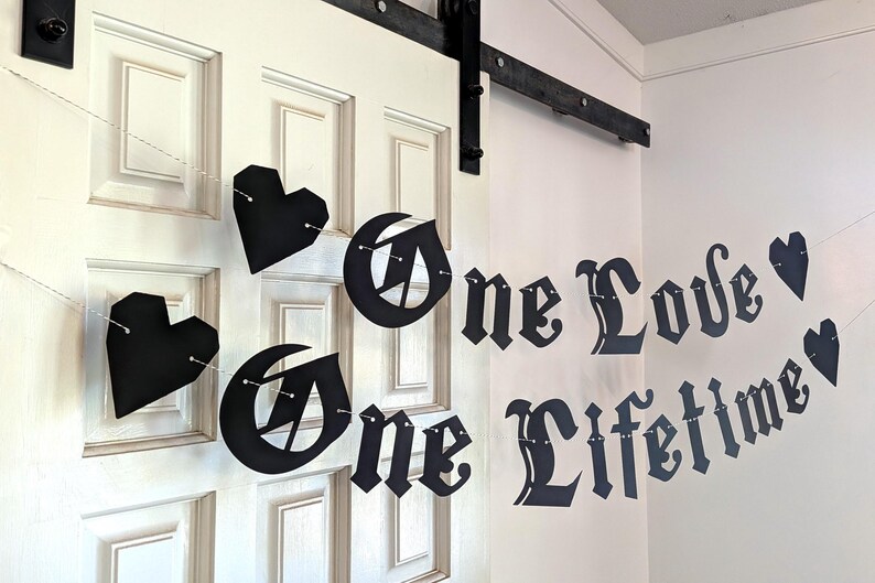 May include: A black paper banner featuring the text One Love One Lifetime in a gothic-style font, interspersed with black heart-shaped cutouts. The individual letters and heart shapes are connected by a thin white string, creating a hanging display.