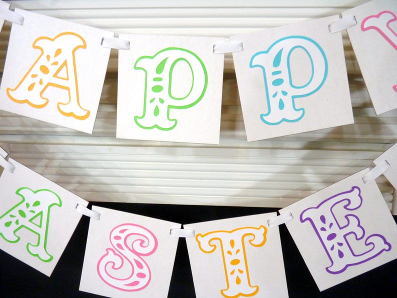 May include: A banner with the words "HAPPY EASTER" spelled out in colorful, decorative letters. The letters are orange, green, blue, pink, and purple, and are attached to a white ribbon.