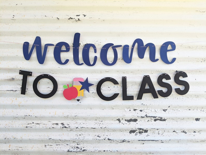 Może przedstawiać: Baner z napisem "welcome TO CLASS" w granatowych i czarnych literach. Baner zawiera czerwone jabłko, ż&oacute;łty oł&oacute;wek i niebieską gwiazdę. Baner jest nawleczony na r&oacute;żowy sznurek.