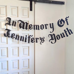 May include: A black banner with the words "In Memory of Jennifer's Youth" in an ornate font hangs in front of a white door. The banner is strung across a white wall and door.