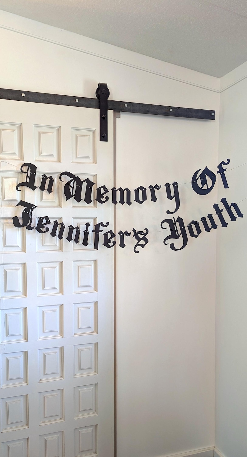 May include: A black banner with the words "In Memory of Jennifer's Youth" in an ornate font hangs in front of a white door. The banner is strung across a white wall and door.