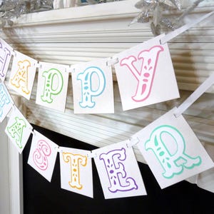 May include: A white banner with the words "HAPPY EASTER" printed in colorful, ornate letters. The letters are in shades of purple, yellow, green, and pink. The banner is strung on a white ribbon, perfect for Easter decorations.