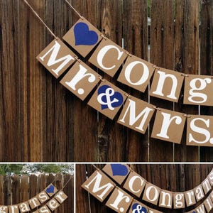 May include: A brown banner with white lettering spelling "Mr. & Mrs. Congrats Wiser" and blue heart accents. The banner is strung on twine and hangs against a wooden fence, perfect for a wedding or anniversary celebration.