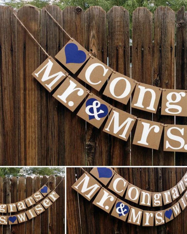 May include: A brown banner with white lettering spelling "Mr. & Mrs. Congrats Wiser" and blue heart accents. The banner is strung on twine and hangs against a wooden fence, perfect for a wedding or anniversary celebration.