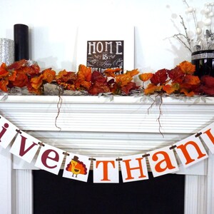 May include: A white mantelpiece decorated for Thanksgiving with a banner reading "GIVE THANKS" in burgundy and orange. The banner includes a turkey illustration. The mantel also features autumn leaves, candles, and a framed sign.
