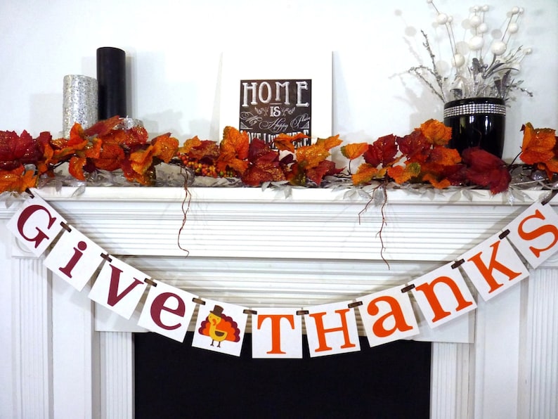 May include: A white mantelpiece decorated for Thanksgiving with a banner reading "GIVE THANKS" in burgundy and orange. The banner includes a turkey illustration. The mantel also features autumn leaves, candles, and a framed sign.