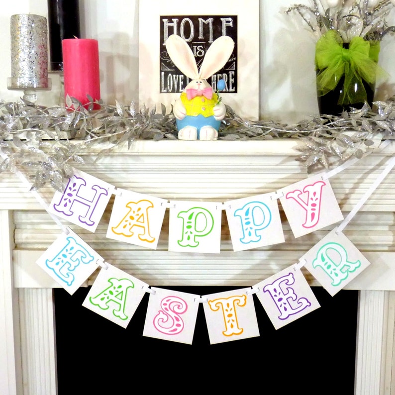 May include: A festive Easter mantel display featuring a colorful "Happy Easter" banner. The banner has multi-colored letters. A bunny figurine, candles, and decorative silver garland complete the spring-themed arrangement.