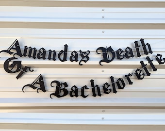 May include: Black metal banner with the text "Amanda's Death or A Bachelorette" in a gothic font, strung on a thin wire. The banner is displayed against a white and silver corrugated metal background.