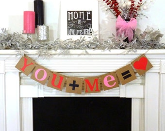 May include: A Valentine's Day mantel display featuring a banner that reads "YOU + ME = ", with a red heart. The mantel also has candles, a framed sign that says "HOME IS HAPPY HERE LOVE LIVES HERE", and decorative floral arrangements.