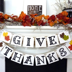 May include: A festive Thanksgiving banner with the words "GIVE THANKS" in black letters. The banner is strung with twine and decorated with colorful leaf cutouts. A fall-themed garland and a framed sign that says "HOME is where the heart is" complete the display.