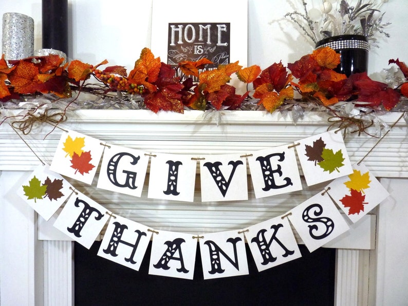 May include: A festive Thanksgiving banner with the words "GIVE THANKS" in black letters. The banner is strung with twine and decorated with colorful leaf cutouts. A fall-themed garland and a framed sign that says "HOME is where the heart is" complete the display.