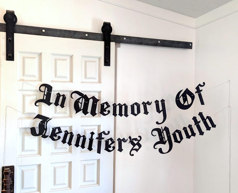 May include: Black banner with the text "In Memory Of Jennifer's Youth" in a gothic font, strung across a white door. The banner is suspended by thin, clear strings.