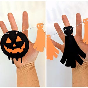 May include: A Halloween garland featuring a black jack-o'-lantern with an orange face and dripping details, along with orange and black paper cutouts strung on a white and black twine. The garland is held up by a hand.