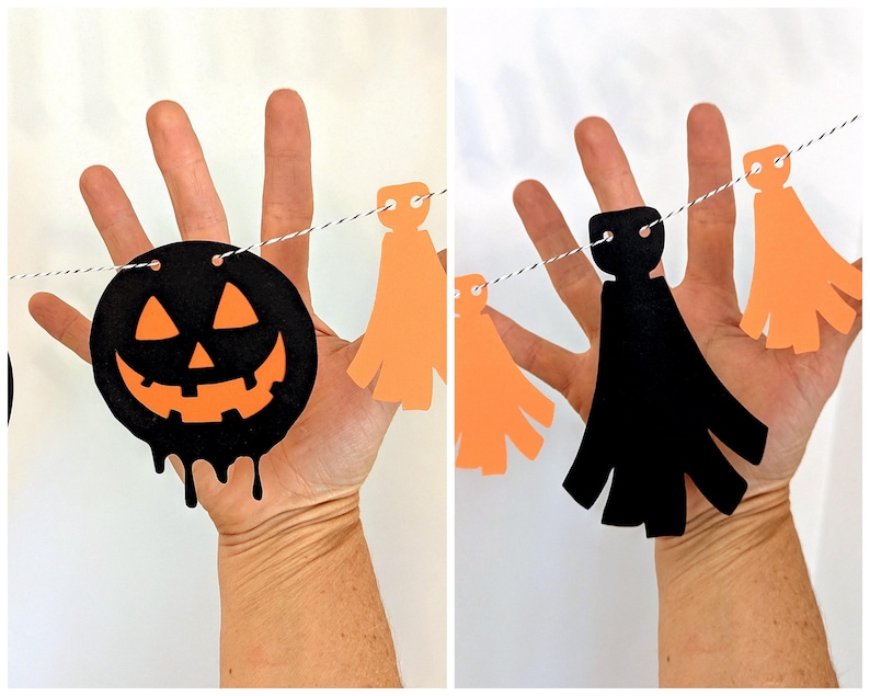 May include: A Halloween garland featuring a black jack-o'-lantern with an orange face and dripping details, along with orange and black paper cutouts strung on a white and black twine. The garland is held up by a hand.