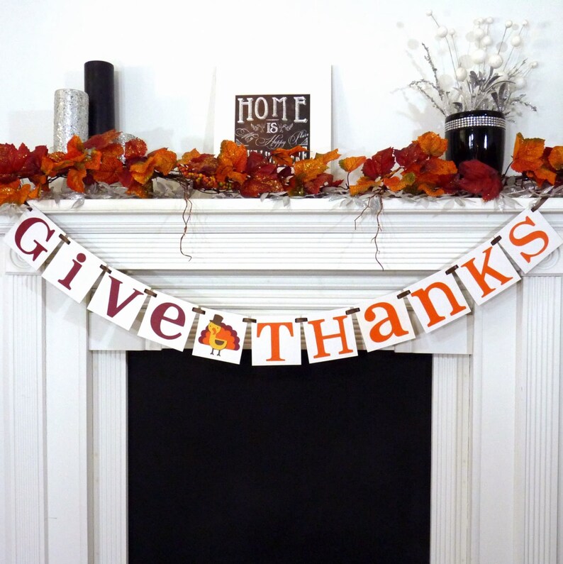 May include: Thanksgiving banner reading "Give Thanks" in burgundy and orange. A turkey illustration is included. The banner is strung across a white mantelpiece decorated with autumn leaves and decorative items.