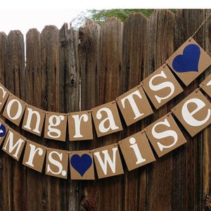 May include: A banner with the words "Congrats Mr. & Mrs. Wiser" in white letters on brown square cards, strung together with twine. Blue hearts accent the design, set against a wooden fence background.