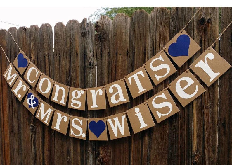 May include: A banner with the words "Congrats Mr. & Mrs. Wiser" in white letters on brown square cards, strung together with twine. Blue hearts accent the design, set against a wooden fence background.