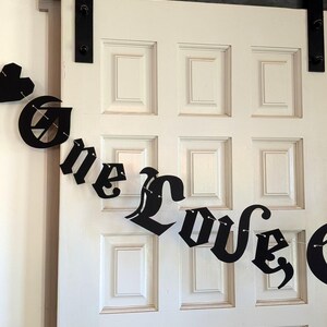 May include: A black decorative banner featuring the text One Love in a gothic-style font, strung on a thin cord. The banner includes a small black heart shape at the beginning and is displayed against a white paneled door with black hardware.