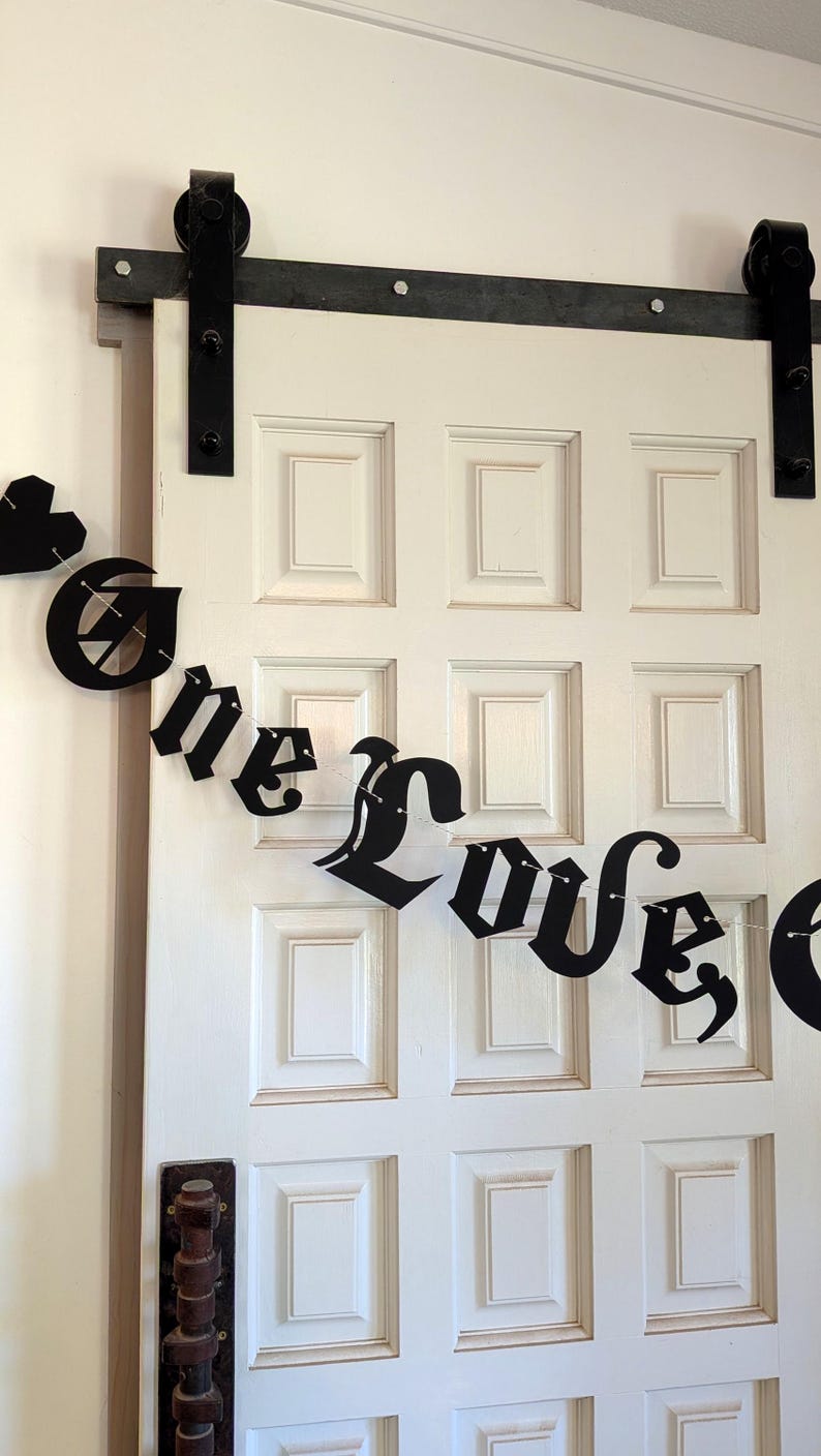 May include: A black decorative banner featuring the text One Love in a gothic-style font, strung on a thin cord. The banner includes a small black heart shape at the beginning and is displayed against a white paneled door with black hardware.