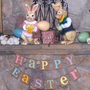 May include: A festive Easter display featuring two rabbit figurines, decorative eggs, and a banner that reads "HAPPY EASTER." The bunnies are in pink and yellow outfits. The display is set against a stone background.