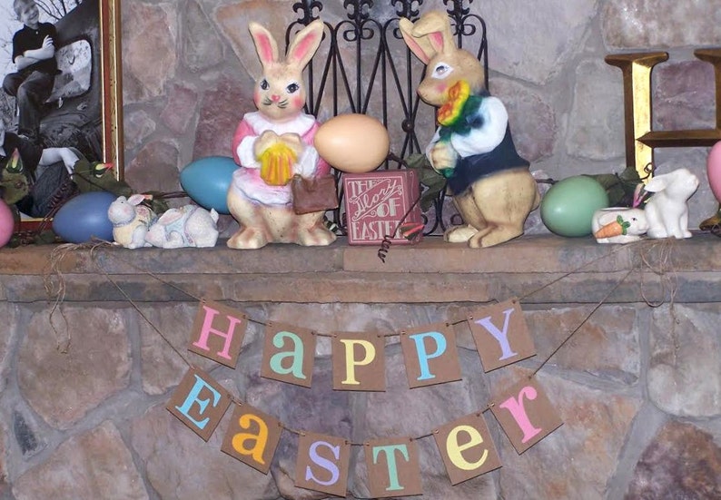 May include: A festive Easter display featuring two rabbit figurines, decorative eggs, and a banner that reads "HAPPY EASTER." The bunnies are in pink and yellow outfits. The display is set against a stone background.