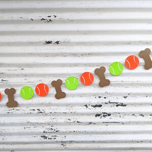 May include: A festive dog-themed banner featuring alternating orange tennis balls, lime green tennis balls, and brown bone shapes. The banner is strung on a white string against a distressed white background.