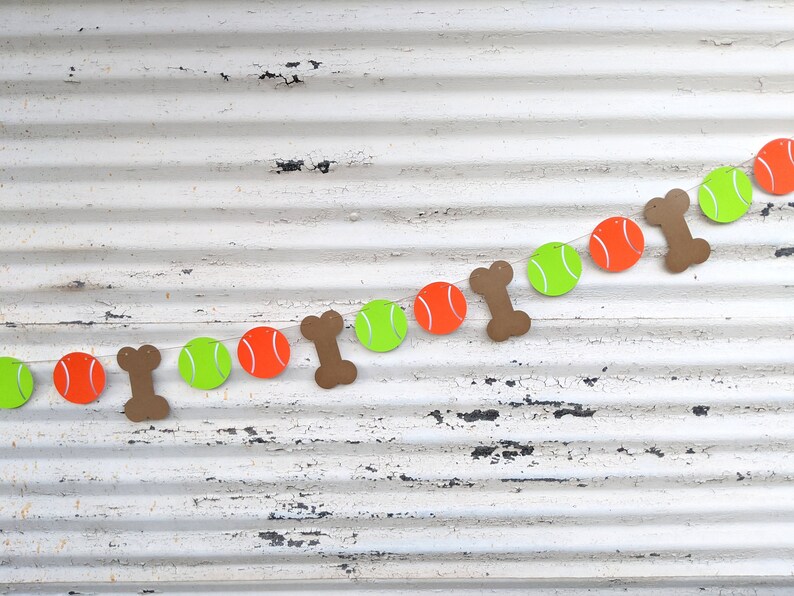 May include: A festive dog-themed banner featuring alternating orange tennis balls, lime green tennis balls, and brown bone shapes. The banner is strung on a white string against a distressed white background.
