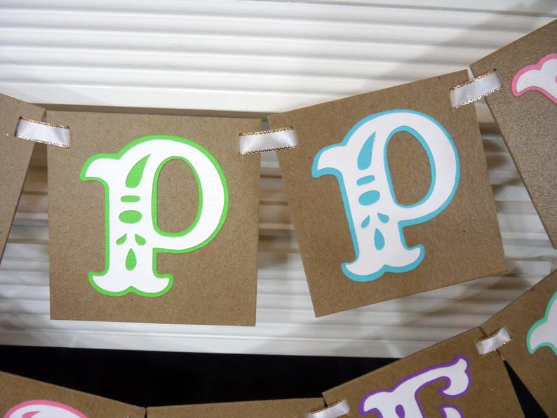 May include: A banner with brown cardstock pennants, each displaying a letter "P". The letters are white with green or blue accents. White ribbon is used to connect the pennants. The banner is likely for a party or celebration.