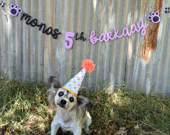 May include: A small dog wearing a party hat with a peach pom-pom. A banner above reads "monas 5th barkday" with paw print accents. The dog is in a grassy area with a corrugated metal background.