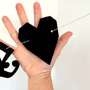 May include: A black geometric heart-shaped cutout made of a flat material, featuring two small holes with a thin black and white twisted string threaded through them, part of a decorative garland string of shapes.