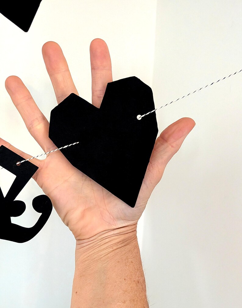 May include: A black geometric heart-shaped cutout made of a flat material, featuring two small holes with a thin black and white twisted string threaded through them, part of a decorative garland string of shapes.