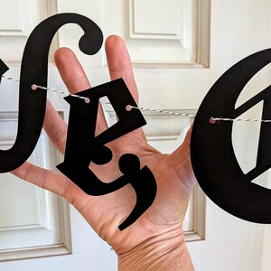 May include: A black letter banner featuring cutout letters strung on a thin black and white twine. The letters are in a decorative script font. The text at the bottom reads: *if you select the 1 strand version, the e in the first Love will come with a comma attached.