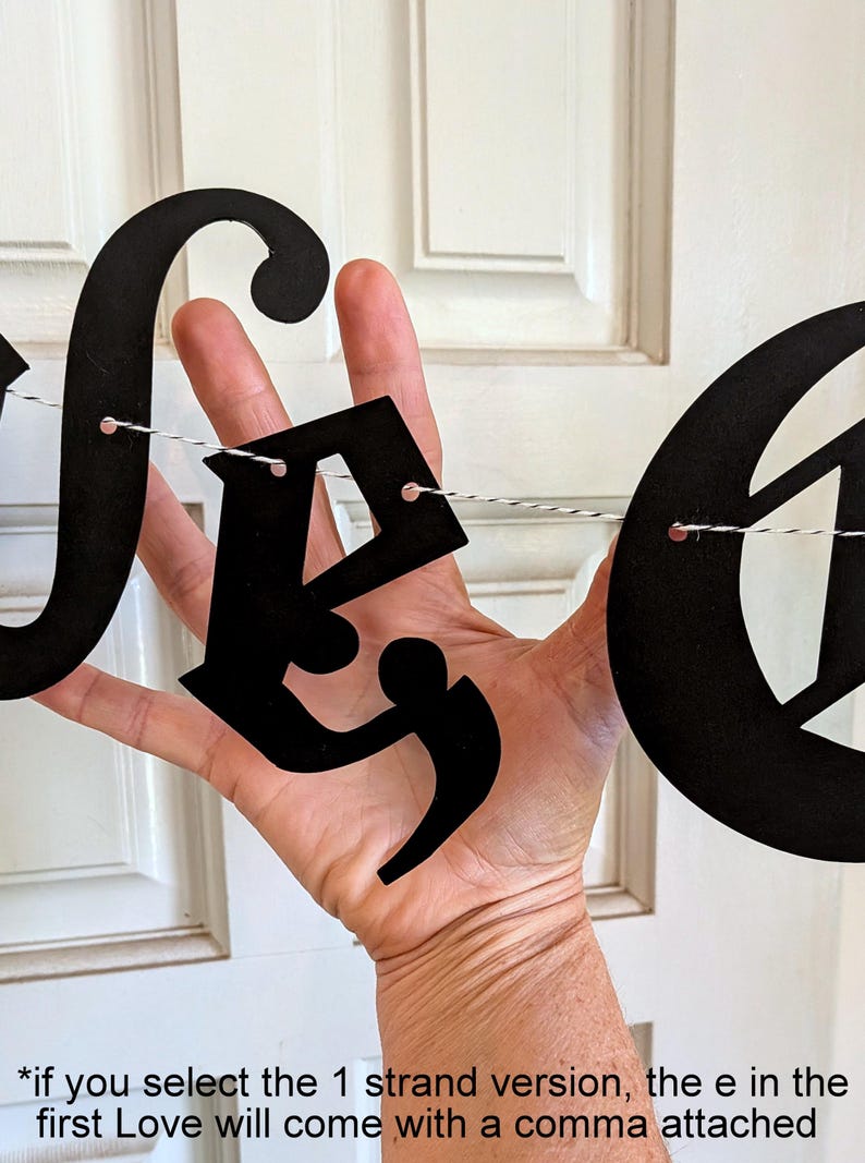 May include: A black letter banner featuring cutout letters strung on a thin black and white twine. The letters are in a decorative script font. The text at the bottom reads: *if you select the 1 strand version, the e in the first Love will come with a comma attached.