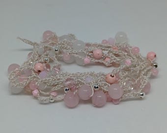Puede incluir: Una pulsera hecha a mano con un delicado diseño de ganchillo en color crema claro. La pulsera está adornada con una variedad de cuentas rosas y transparentes, incluyendo cuentas redondas más grandes y cuentas de acento más pequeñas. Cuentas de metal plateado están intercaladas.