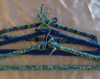 Puede incluir: Tres perchas para ropa cubiertas con hilo de ganchillo. La percha superior presenta un hilo jaspeado en azul, verde y morado. La percha central es azul sólido y la inferior es de hilo jaspeado en azul claro, verde y amarillo.