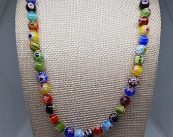 Puede incluir: Un collar de cuentas de colores con una variedad de cuentas de vidrio en rojo, azul, amarillo, verde y negro. El collar tiene un colgante plateado con un diseño circular. Se muestra en un maniquí.