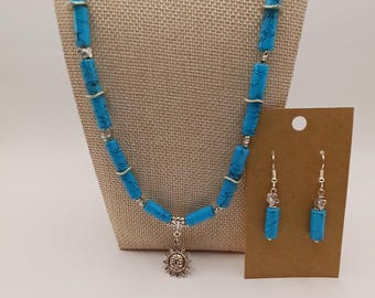 Puede incluir: Un conjunto de collar y pendientes con cuentas cilíndricas de color azul brillante con vetas oscuras, acentuadas por pequeños espaciadores plateados. El collar incluye un colgante de sol plateado y pendientes a juego con cuentas.