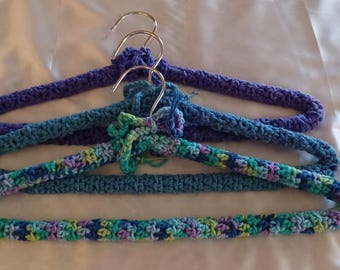 Puede incluir: Tres perchas para ropa cubiertas con hilo tejido a ganchillo a mano. La percha superior es de color morado sólido, la del medio es de color verde azulado sólido y la inferior presenta un patrón de hilo jaspeado en tonos azul, verde, morado y amarillo. Cada percha tiene un gancho de metal.