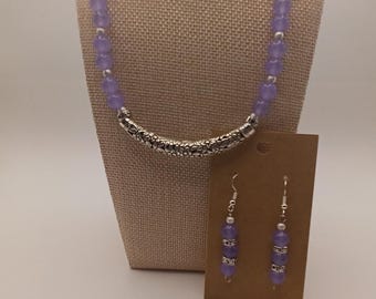 Puede incluir: Un conjunto de joyería compuesto por un collar y pendientes a juego. El collar presenta cuentas redondas de color morado translúcido intercaladas con pequeños espaciadores plateados y una pieza central curva y grabada. Los pendientes incluyen cuentas moradas y detalles plateados en ganchos.