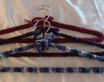 Puede incluir: Tres perchas para ropa cubiertas con hilo tejido a mano. La percha trasera es de color rojo oscuro sólido, la del medio es de color morado oscuro y la delantera presenta un patrón variado de hilo azul, morado y blanco. Cada una tiene un gancho de metal plateado.