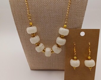 Puede incluir: Un collar de cadena en tono dorado con seis cuentas blancas opacas y lisas en forma de barril, separadas por pequeñas cuentas espaciadoras doradas, junto con un par de pendientes a juego con cuentas blancas en ganchos dorados.
