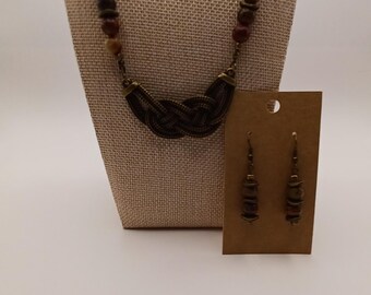 Puede incluir: Un conjunto de collar y pendientes con cuentas en tonos marrones y oscuros, con un colgante central de metal color bronce en forma de nudo. El collar tiene una cadena de cuentas y los pendientes a juego se presentan en una tarjeta marrón.