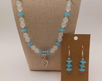 Puede incluir: Un conjunto de collar y pendientes con cuentas blancas translúcidas y cuentas de disco de color azul turquesa brillante. El collar incluye un colgante de metal plateado en forma de corazón con un diseño recortado. Los pendientes son colgantes.