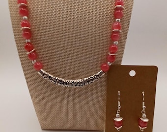 Puede incluir: Un conjunto de collar y pendientes con cuentas redondas de color rosa translúcido intercaladas con pequeños espaciadores de tono plateado. El collar incluye un tubo central de metal plateado curvado con un patrón floral en relieve. Los pendientes a juego presentan una cuenta rosa y detalles plateados.