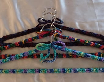 Puede incluir: Tres perchas para ropa cubiertas con hilo de ganchillo. La percha superior es de color azul marino oscuro, la del medio presenta un patrón de hilo multicolor y la inferior está cubierta con hilo jaspeado en tonos turquesa, azul y violeta.